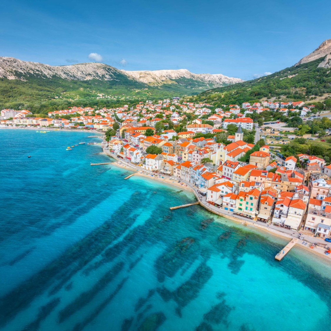 Aerial view of buildings with orange roofs, beach, mountains, blue sea in summer day. Vacation in beautiful Baska city, Krk island, Croatia. Top view of houses, streets, promenade, adriatic sea bay
